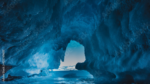 An unusual grotto in the rock is completely covered with shiny blue ice, icicles. In the gap of the entrance you can see the azure sky, snow-capped mountains. Baikal