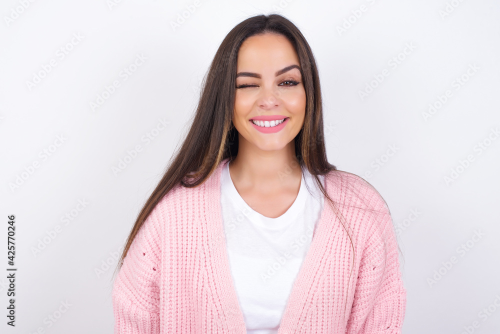 Coquettish Young beautiful caucasian woman wearing pink knitted jacket over white wall smiling happily, blinking at camera in a playful manner, flirting with you. Coquettish Young beautiful caucasian woman wearing pink knitted jacket over white wall smiling happily, blinking at camera in a playful manner, flirting with you.