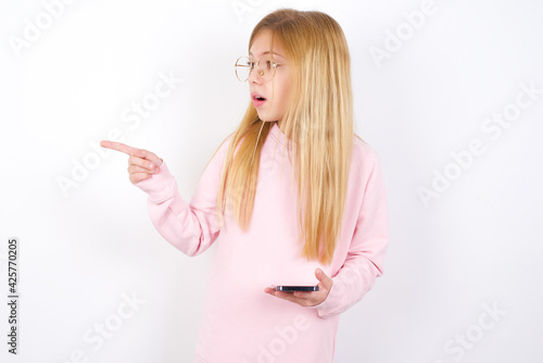 Stunned beautiful caucasian little girl wearing pink sweater points sideways right copy space, recommends product, sees astonishing thing