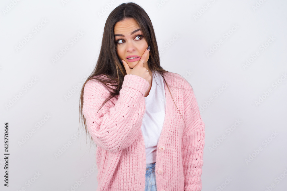 Young beautiful caucasian woman wearing pink knitted jacket over white wall Thinking worried about a question, concerned and nervous with hand on chin. Young beautiful caucasian woman wearing pink knitted jacket over white wall Thinking worried about a question, concerned and nervous with hand on chin.