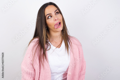 Schilderij op canvas Young beautiful caucasian woman wearing pink knitted jacket over white wall showing grimace face crossing eyes and showing tongue