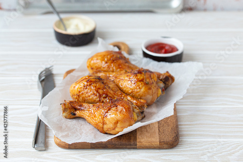 Roasted golden chicken legs drumsticks on baking paper on wooden board served with ketchup and mayonnaise on rustic white wooden table, angle view 
