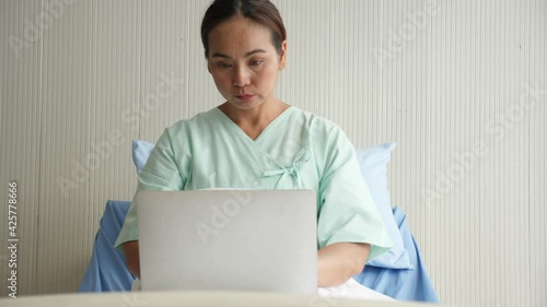 Medium shot portrait of middle-gaed adult Asian woman wearing patient outfit, sitting on bed, using laptop computer, working in hospital ward. Busy life of employee duting sick leave concept.