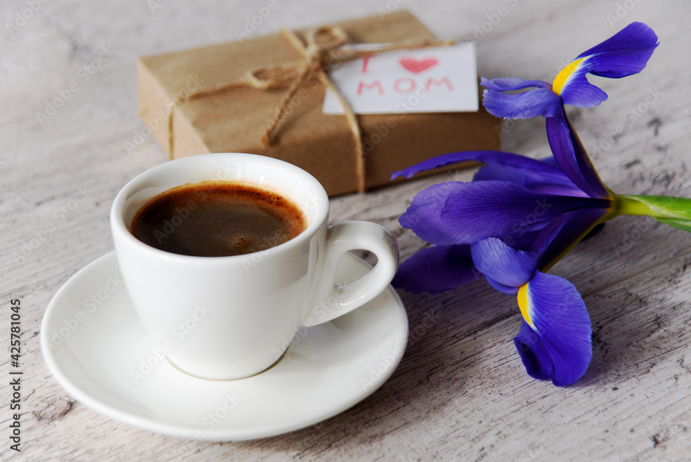 White cup of coffee, gift box with message for mom and iris flower on gray wood table. Mother's Day holiday concept.