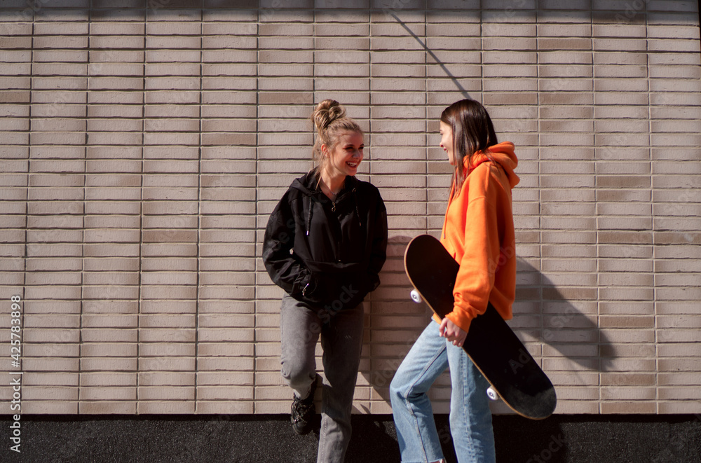 Two teenage skater girls are hanging out in the neighborhood, chatting and smiling.