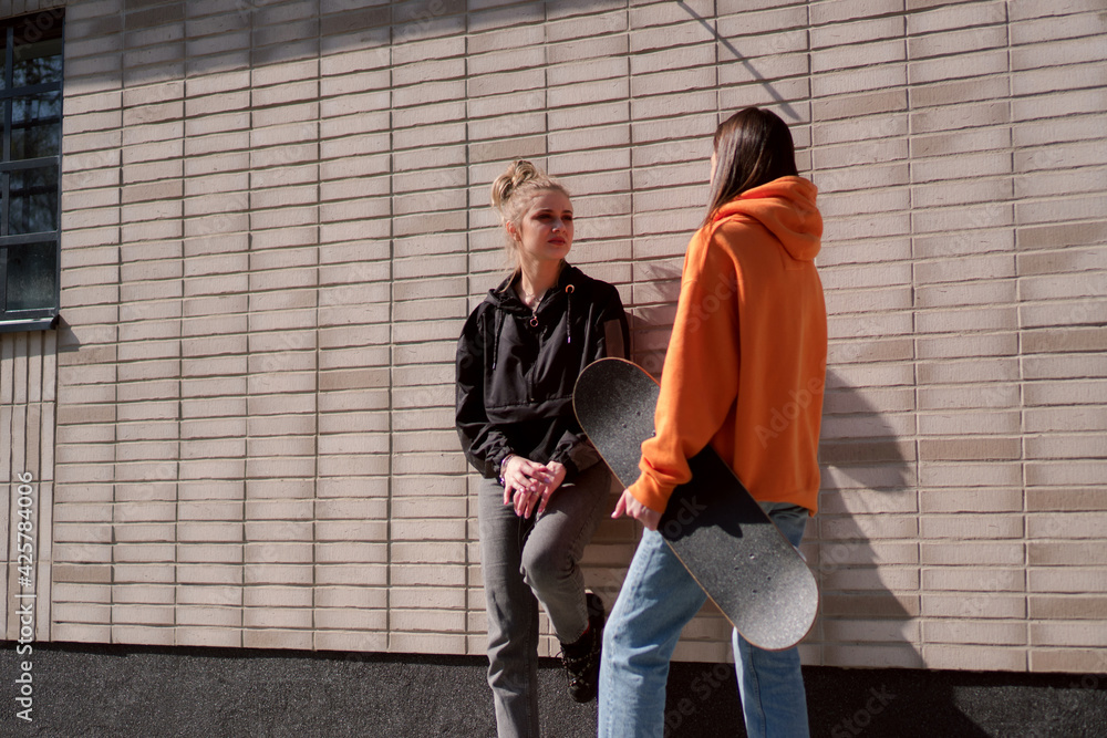 Two teenage skater girls are hanging out in the neighborhood, chatting and smiling.