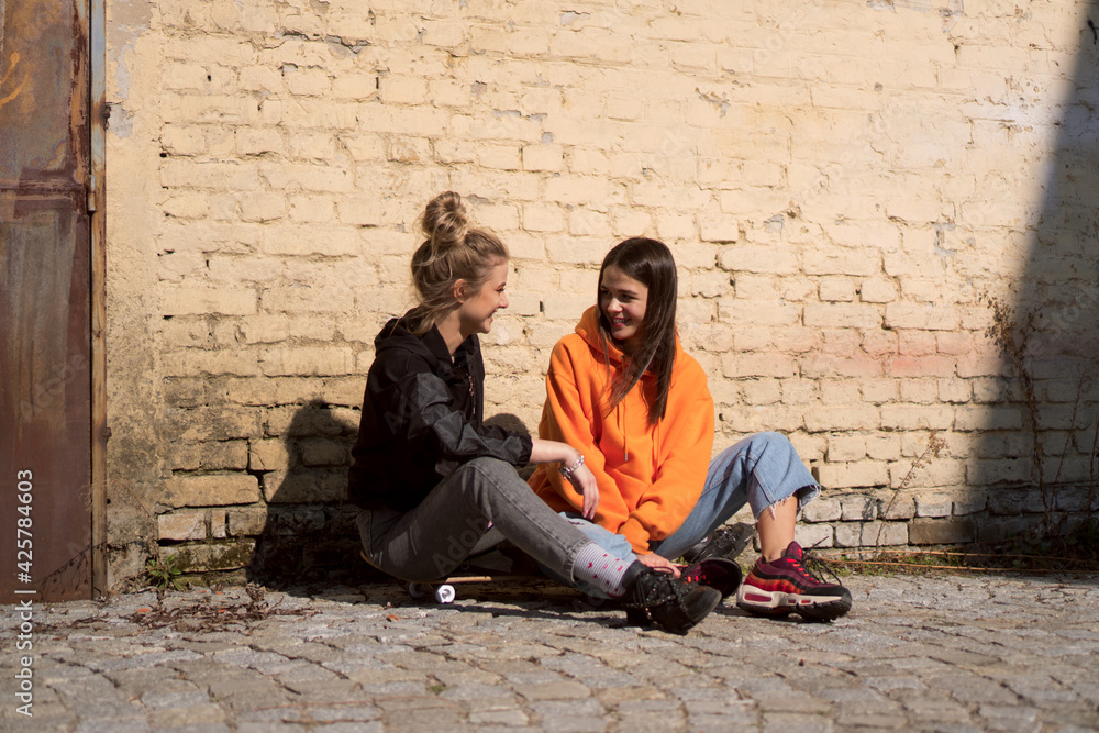 Two teenage skater girls are hanging out in the neighborhood, chatting and smiling.