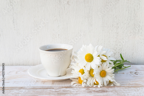 White vintage cup of coffee or tea and bouquet of white daisies on white paint wooden background; space for text