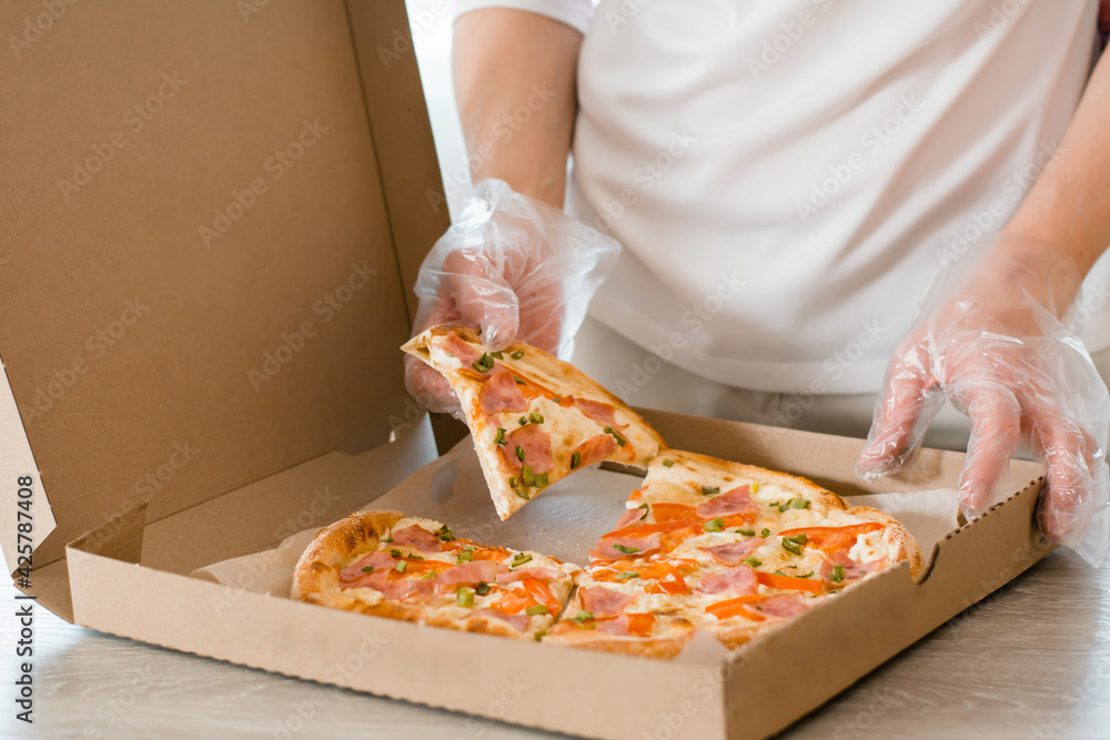Takeaway  food. A woman in disposable gloves takes a slice of pizza from a cardboard box on the table in the kitchen.