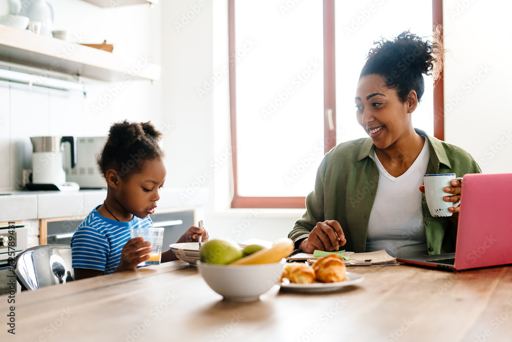 Black smiling woman working while having breakfast with her daughter