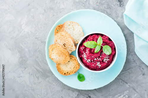 Homemade baked beetroot hummus in a bowl with sesame seeds and basil and baked small toast on a plate on the table. Top view