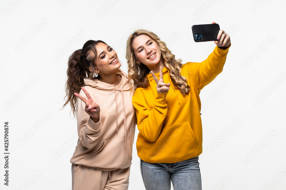 Portrait of a two women making selfie photo on smartphone while showing two fingers sign on gray background Portrait of a two women making selfie photo on smartphone while showing two fingers sign on gray background