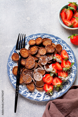 Mini chocolate pancake cereal with strawberries for breakfast on gray concrete table. Trendy home breakfast with tiny pancakes. Top view