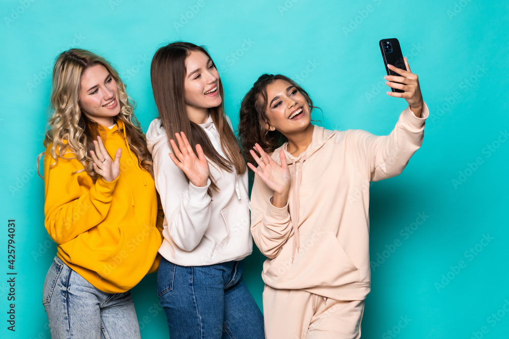 Young three multiethnic women friends make video call on phone posing isolated over turquoise background Young three multiethnic women friends make video call on phone posing isolated over turquoise background