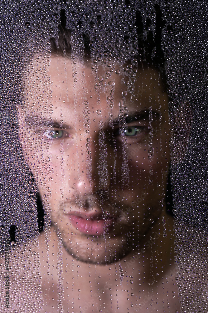 Handsome man with green eyes looking out from behind rainy glass or man in shower cubicle 
