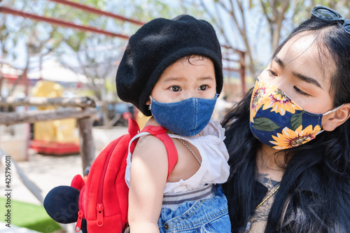 Little child girl and Asian mother wearing medical protective mask and holding her daughter in a crowded public place in park or tourist. Happy baby and mom family playing outside Covid-19 pandemic.