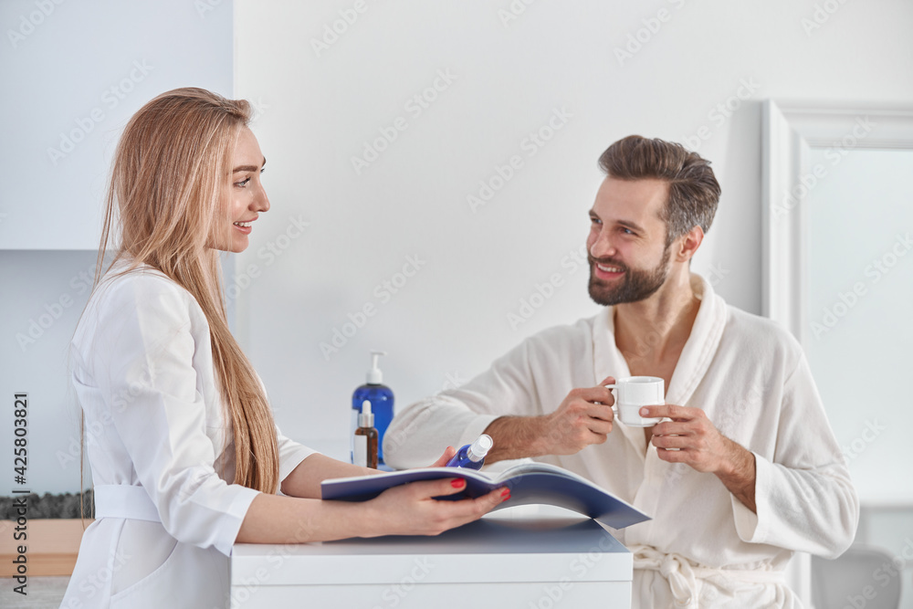 Obraz premium Medical worker consults with brochure and beauty treatments to patient. Patient young man standing near the reception and holding tea