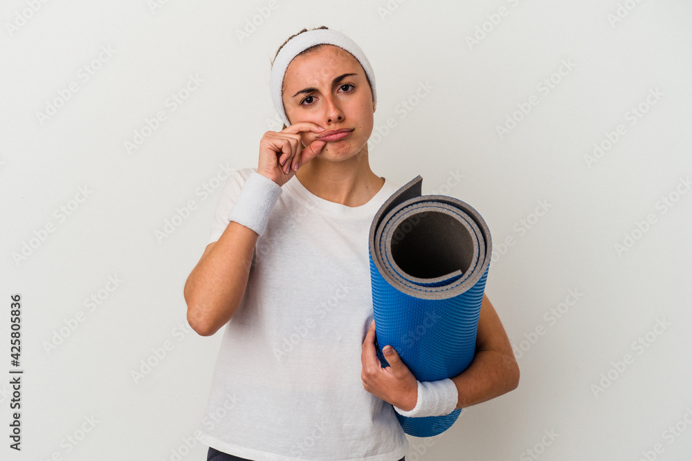 Young sporty woman holding a mat isolated on white background with fingers on lips keeping a secret. Young sporty woman holding a mat isolated on white background with fingers on lips keeping a secret.