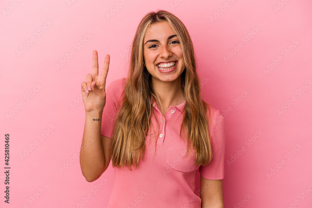 Young blonde caucasian woman isolated on pink background showing victory sign and smiling broadly. Young blonde caucasian woman isolated on pink background showing victory sign and smiling broadly.