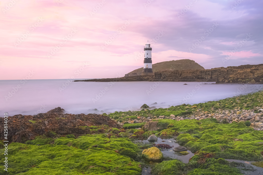 Colourful idyllic pink sky sunset or sunrise seascape at Penmon Point and Trywn Du Lighthouse along the green algae rocky shoreline in Anglesey, North Wales, UK.