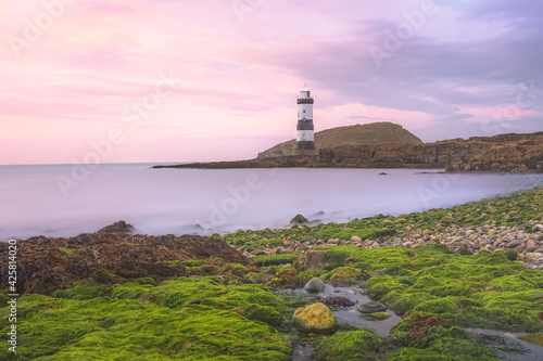 Colourful idyllic pink sky sunset or sunrise seascape at Penmon Point and Trywn Du Lighthouse along the green algae rocky shoreline in Anglesey, North Wales, UK.