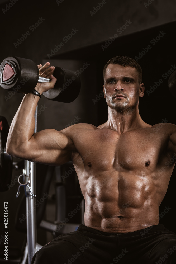 Fitness in gym, sport and healthy lifestyle concept. Handsome athletic man with naked torso making exercises. Bodybuilder male model training muscles lifting large dumbbells up
