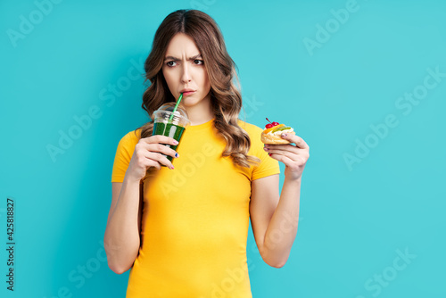 Unhappy woman on dieting drinking detox juice holding cake in hand choosing healthy food