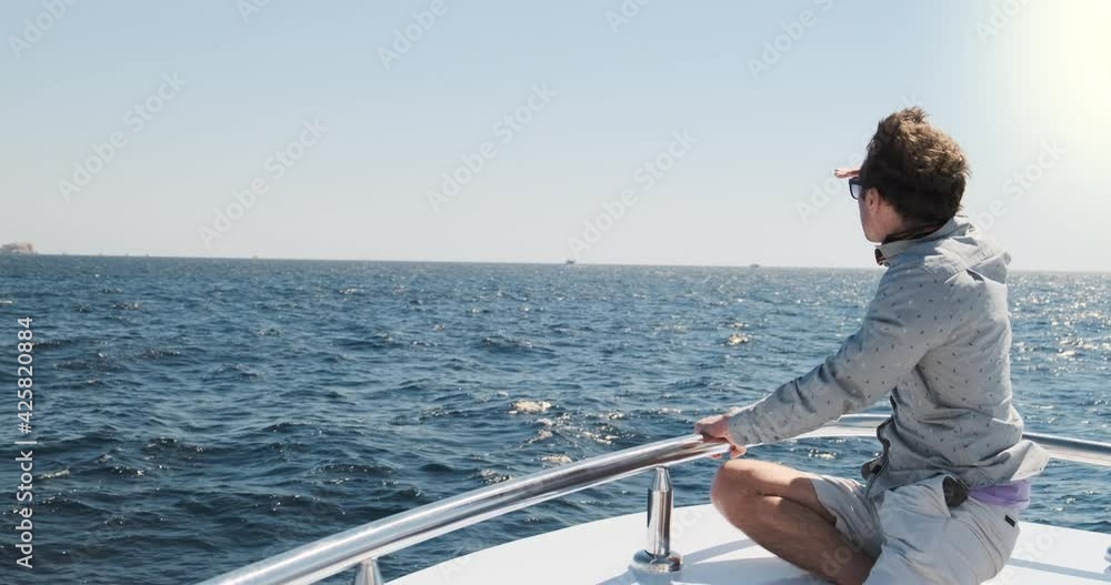 Man on yacht looks in distance, sunny morning. White sailing boat on blue sea waves, handsome man in light clothes, sunglasses sitting on bow. Luxury voyage summer travel adventure. Nautical lifestyle Man on yacht looks in distance, sunny morning. White sailing boat on blue sea waves, handsome man in light clothes, sunglasses sitting on bow. Luxury voyage summer travel adventure. Nautical lifestyle
