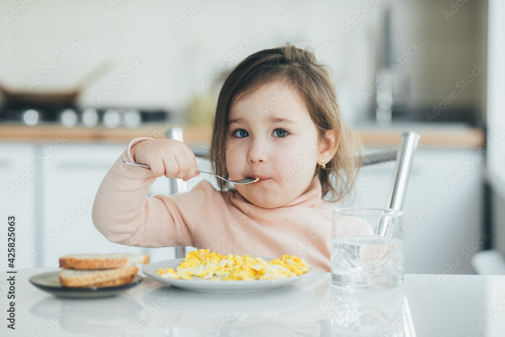 Baby girl in the afternoon in the white light kitchen in a pink sweater eating an omelet Baby girl in the afternoon in the white light kitchen in a pink sweater eating an omelet