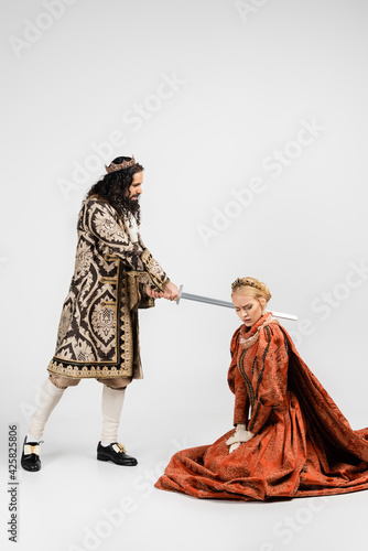 full length of cruel hispanic king in medieval clothing holding sword near scared blonde queen in crown sitting on white