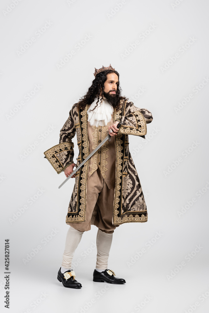 full length of hispanic king in medieval clothing and crown holding sword on white