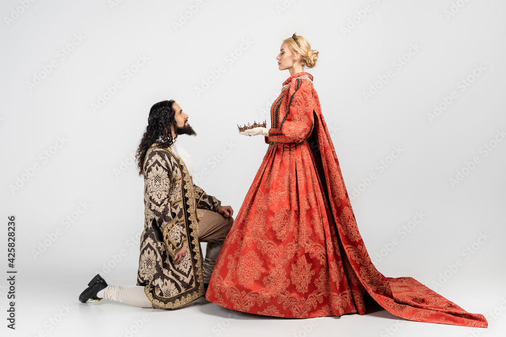 side view of blonde queen holding crown near hispanic king in medieval clothing standing on knee on white