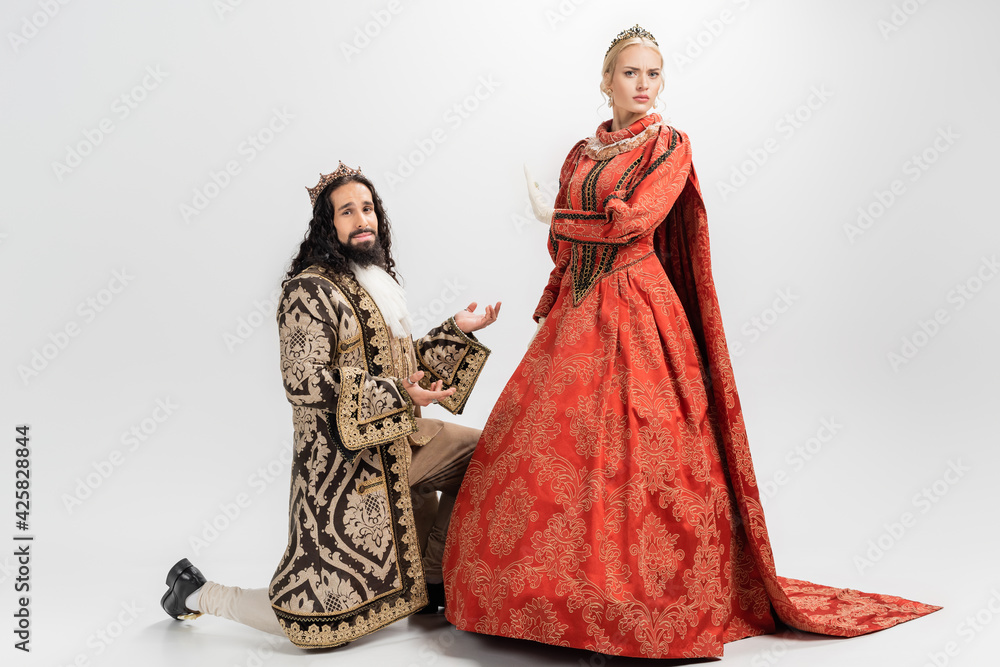hispanic king in crown and medieval clothing standing on knee while blonde queen showing no gesture on white