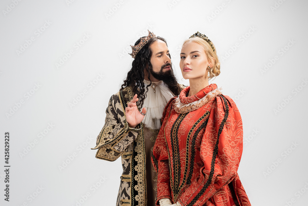 hispanic king in crown and medieval clothing looking at blonde wife isolated on white