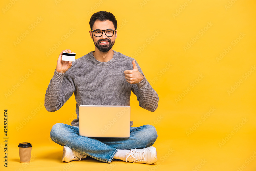 Full length of a cheerful young man sitting with legs crossed isolated over yellow background, using laptop computer, showing credit card. Full length of a cheerful young man sitting with legs crossed isolated over yellow background, using laptop computer, showing credit card.