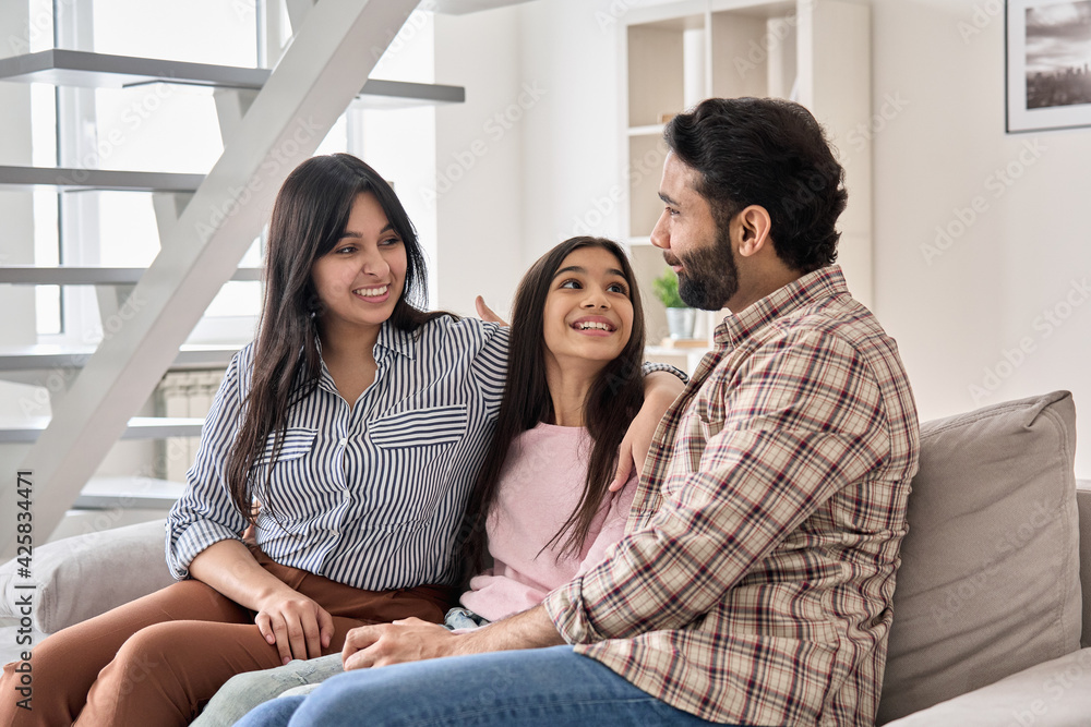 Happy indian family couple with teen child daughter bonding, hugging, talking sitting on couch at home. Smiling husband and wife embracing child spending time together relaxing on sofa in living room Happy indian family couple with teen child daughter bonding, hugging, talking sitting on couch at home. Smiling husband and wife embracing child spending time together relaxing on sofa in living room