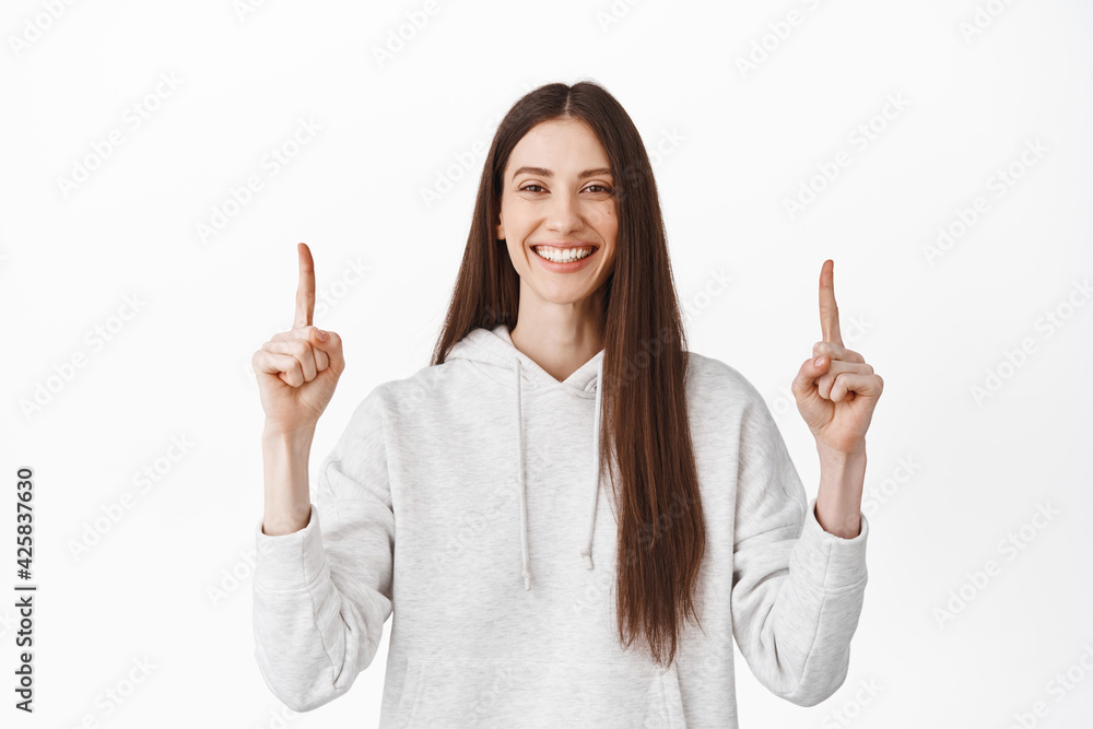 Confident smiling woman recommending logo, click on website banner, pointing fingers up and looking at camera, showing best deal in store, standing over white background Confident smiling woman recommending logo, click on website banner, pointing fingers up and looking at camera, showing best deal in store, standing over white background