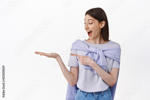 Hey whats it in my head. Amazed girl lookg at her open palm and pointing empty object, showing promo product, standing excited against white background photo