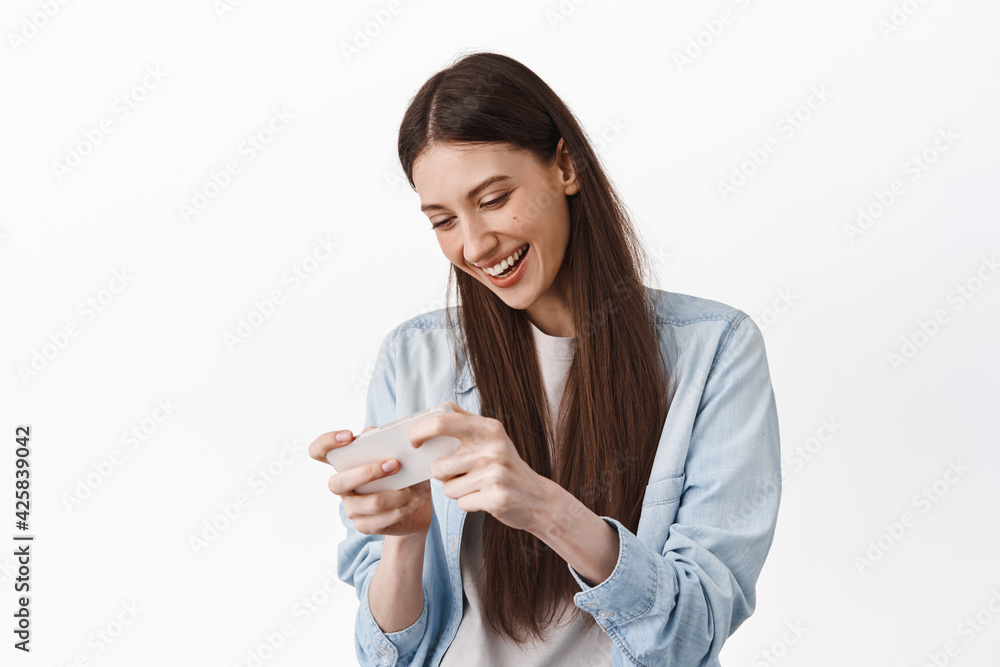 Happy young woman playing video game on smartphone, laughing and looking at phone screen, enjoying gaming, standing against white background Happy young woman playing video game on smartphone, laughing and looking at phone screen, enjoying gaming, standing against white background