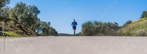 A young white european and athletic man is running in the countryside on a sunny day. The guy is wearing blue clothes and a cap.