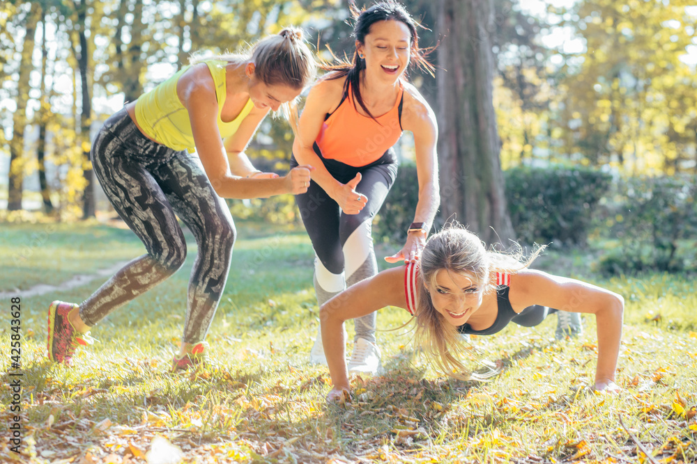 Fototapeta premium Fitness group working out at park. Team of three slim women outdoor doing fitness exercise. Laughing girls counting for their friend during plank exercise in park.