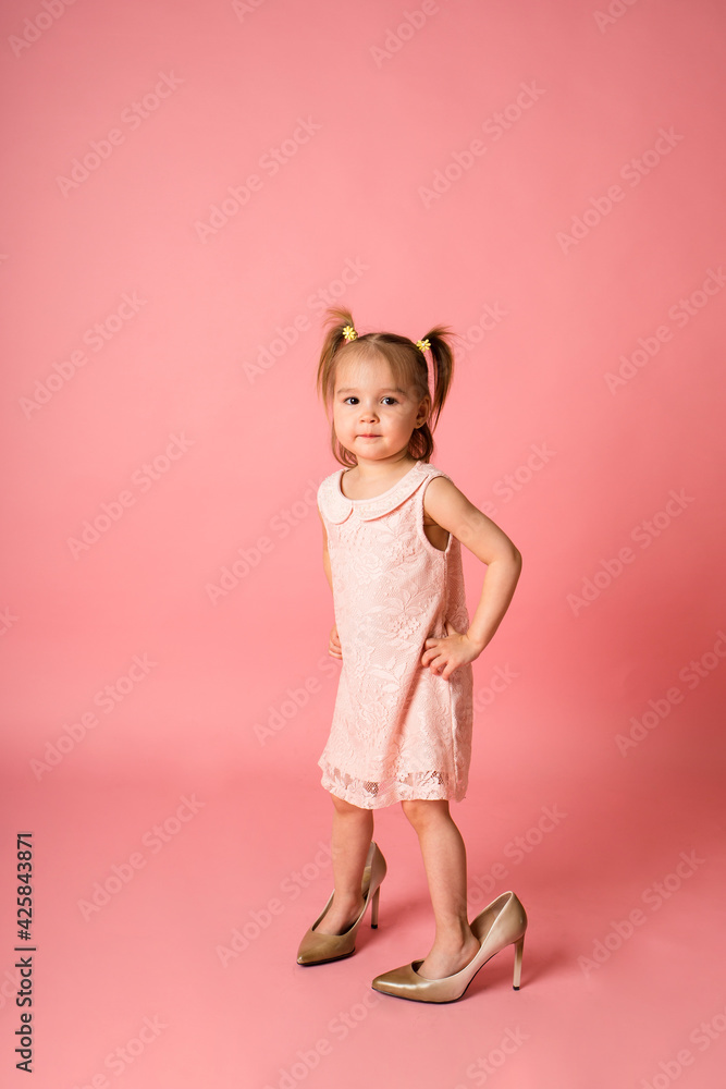 little girl in a pink lace dress and her mother's shoes stands on a pink background with space for text