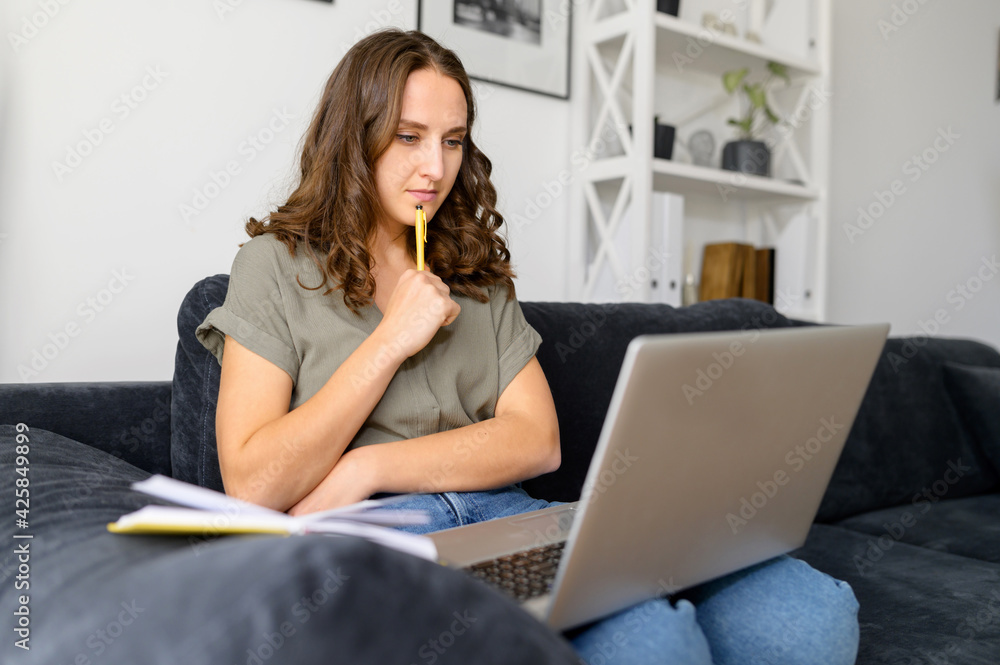 Thoughtful young woman with a laptop, looking on the screen concentrated, takes a notes. Female student studying on the distance, watching online webinars, video lectures sitting on the couch at home Thoughtful young woman with a laptop, looking on the screen concentrated, takes a notes. Female student studying on the distance, watching online webinars, video lectures sitting on the couch at home