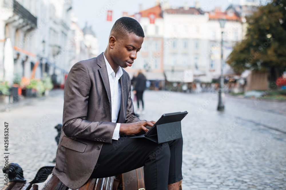 Focused black man in stylish suit sitting on wooden bench outdoors and working on digital tablet. Handsome businessman typing on modern gadget on city street. Focused black man in stylish suit sitting on wooden bench outdoors and working on digital tablet. Handsome businessman typing on modern gadget on city street.