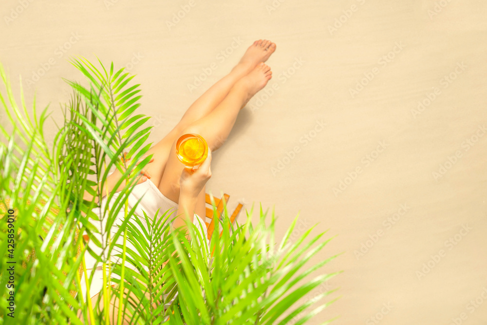 Fototapeta premium Alone woman sitting under palm tree branches with glass of water with slice orange. Female relaxation on the sand of the beach at summer vacation. Top view
