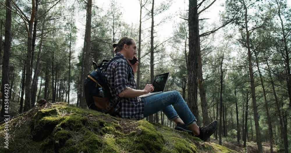 Young hipster man using laptop in forest and talking on the phone, freelance working in nature, fast 5g internet connection. Young hipster man using laptop in forest and talking on the phone, freelance working in nature, fast 5g internet connection.
