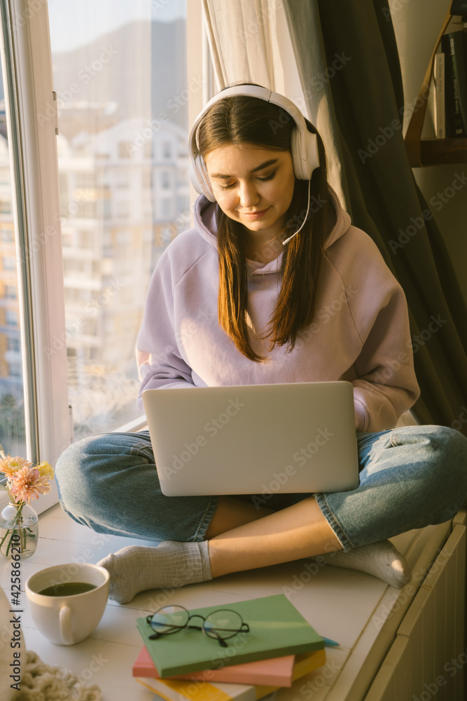Fototapeta premium Smiling woman with headphones working online with laptop. Girl freelancer sitting near window on the sunset at home. Distance learning and remote job concept.