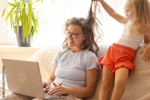 Distant home office during lockdown.Little daughter bothering interfering mother freelancer working using laptop, holding video call with clients, sitting on sofa indoor.Playful girl pulling mom hair