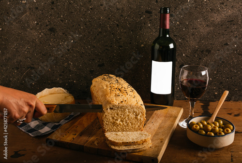 Homemade and artisan bread of morita chili and spices in a stick, a person with a knife cutting it into slices on a wooden table. Accompanied by olives, cheese and red wine. photo