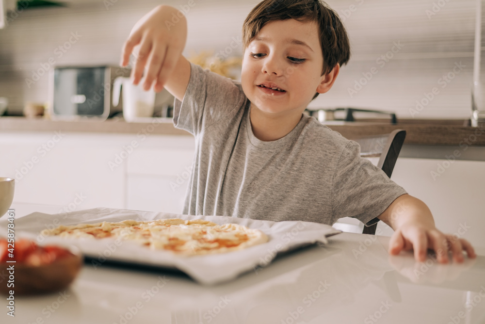 Little kid boy making pizza sitting at the table on the kitchen. Children helping in cooking lifestyle image Little kid boy making pizza sitting at the table on the kitchen. Children helping in cooking lifestyle image